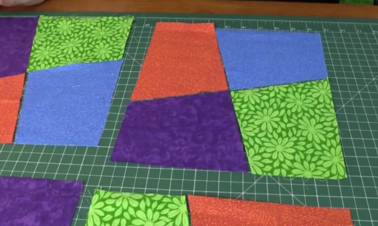 Colorful fabric pieces, including green floral, orange textured, blue swirling, and purple swirling patterns, are laid out on a green cutting mat with grid lines, forming sections of a sewing or quilting project.