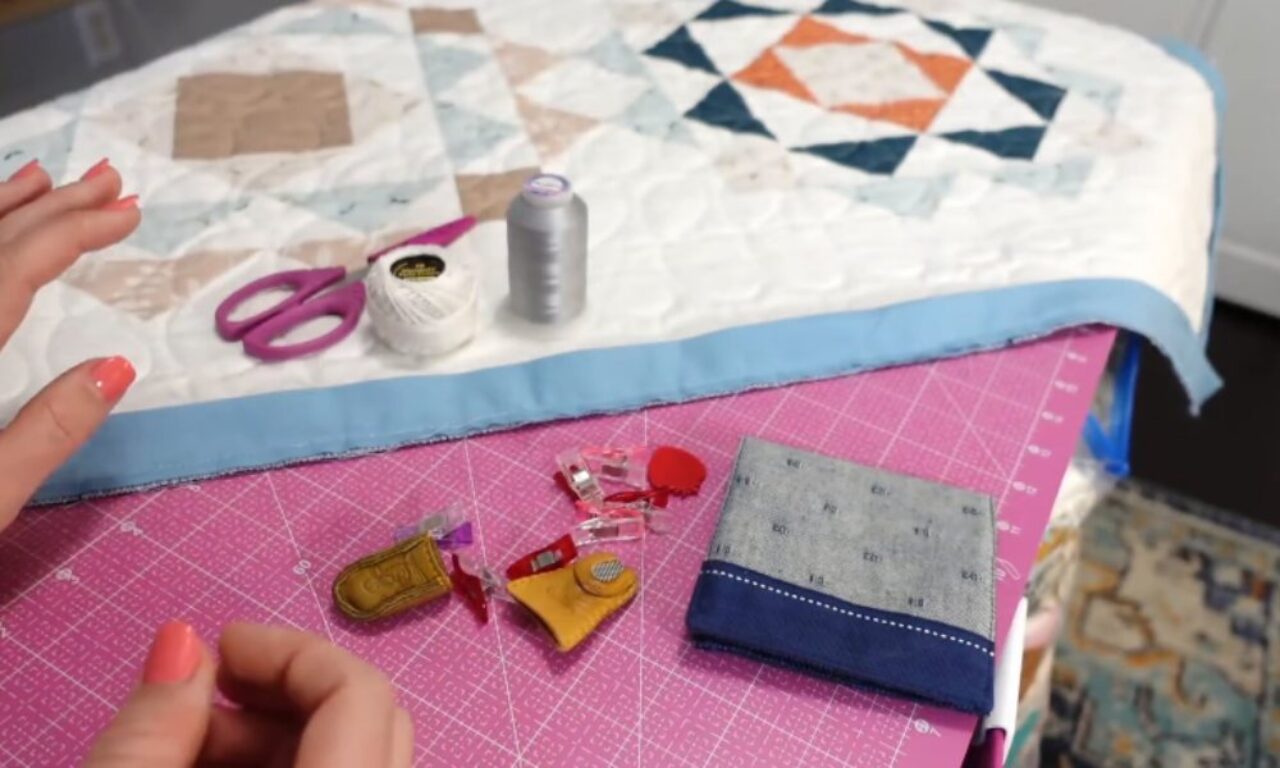A quilted project with geometric patterns and blue binding is shown with sewing supplies including scissors, thread, clips, and a thimble on a pink cutting mat.
