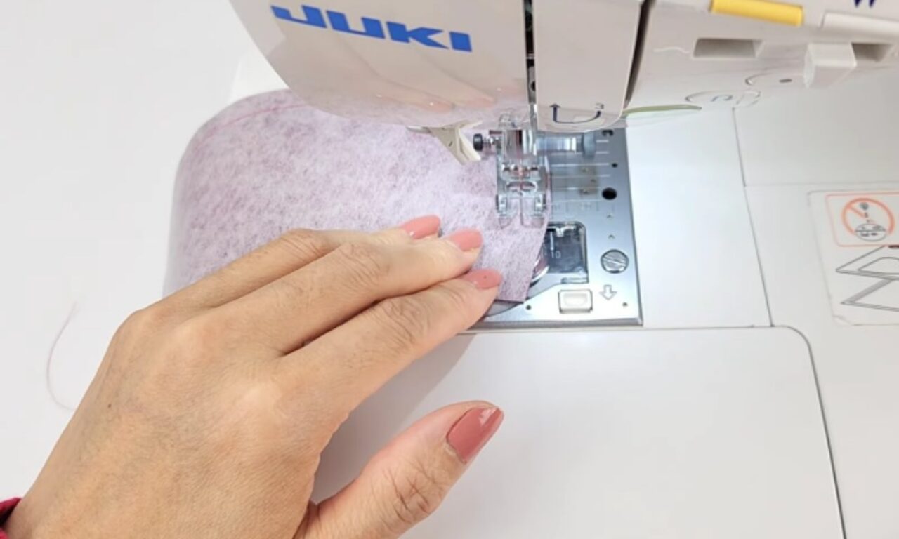 a person's hand guiding a piece of light purple fabric under the needle of a white Juki sewing machine. The person's nails are neatly manicured with light pink polish, and the sewing machine is in the process of stitching the fabric. The work area is clean and well-lit.