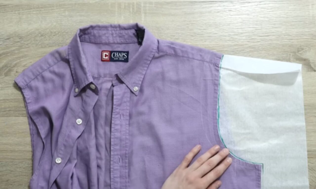 A lavender button-up shirt with the right sleeve removed is laid flat on a light wooden surface. A piece of white paper or fabric, shaped like a sleeve pattern, is placed on the right armhole. A hand is holding down the pattern piece, suggesting a sewing or upcycling project.