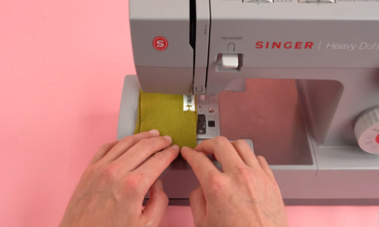 A person is using a Singer Heavy Duty sewing machine to sew a piece of green fabric. The hands are carefully guiding the fabric through the machine, which is set against a pink background. The sewing machine features a red "S" logo and various control buttons and dials.