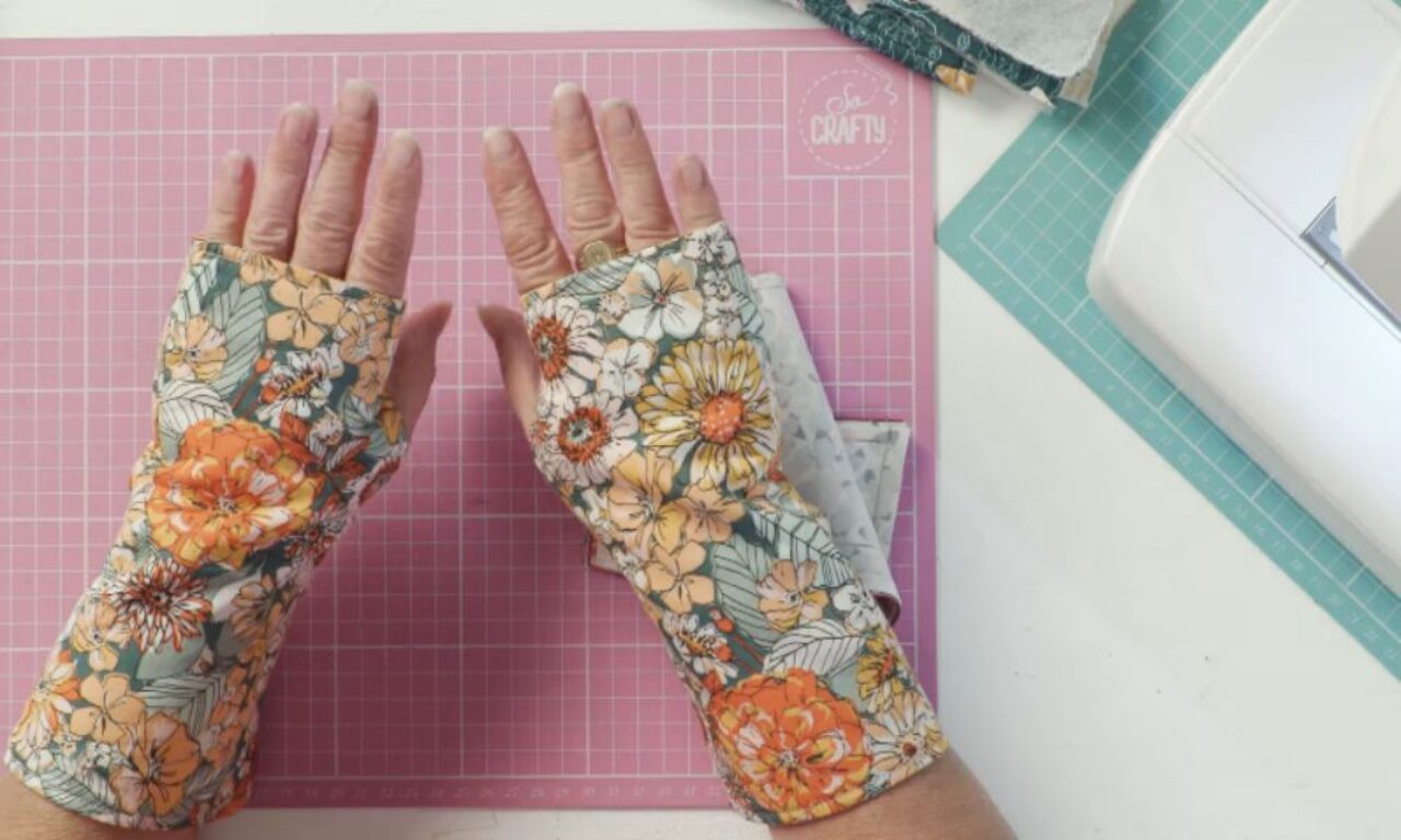 A pair of hands, palms facing up, models handmade fingerless gloves. The gloves feature a vibrant floral pattern with orange, yellow, and white flowers on a muted teal background, and extend up the forearms. They are displayed over a pink gridded cutting mat, with a sewing machine and fabric scraps visible in a crafting environment.