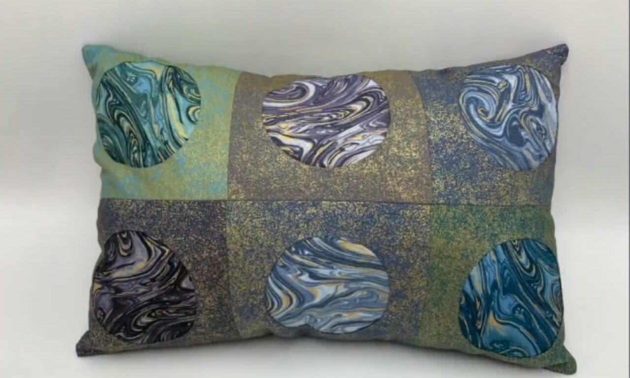 A rectangular throw pillow with a patchwork design of six blocks in muted green, blue, and grey/gold speckled fabric, each featuring a large circular appliqué with a swirling marbled pattern in shades of blue, green, purple, grey, and yellow.