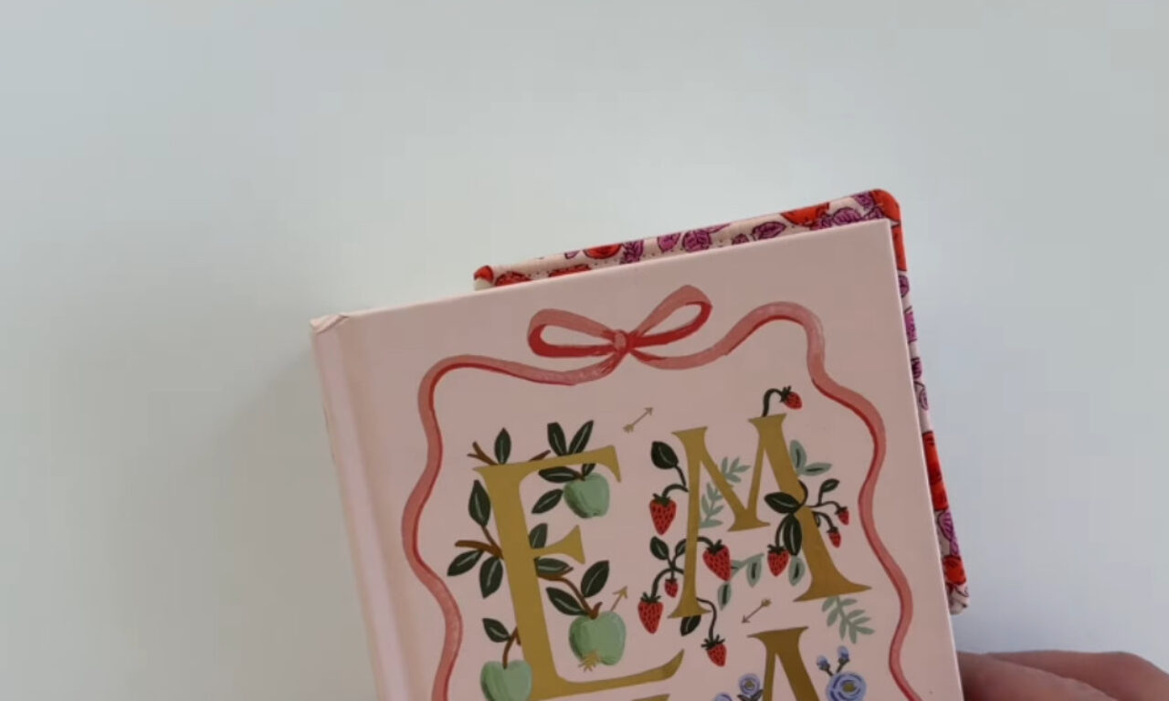 The cover features elegant, gold-colored letters surrounded by decorative elements such as green leaves, red strawberries, and blue flowers. A pink ribbon is tied around the top of the cover, adding a charming, vintage touch. The background of the cover is light pink, and the book is held at an angle against a plain white backdrop.