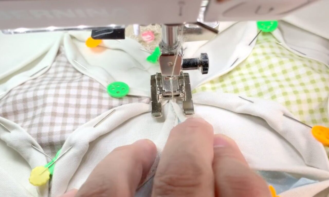A close-up view of a sewing machine foot pressing down on a piece of fabric with colorful pins scattered around it. The fabric features a checkered pattern with white, green, and brown squares. A hand is visible holding the fabric in place, and the sewing machine needle is poised to make a stitch.