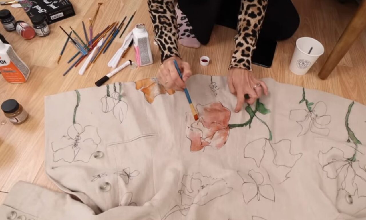 A person wearing leopard print sleeves is painting floral designs on a light-colored fabric spread out on a wooden floor. Various art supplies, including paintbrushes, paints, and a palette, are scattered around them.