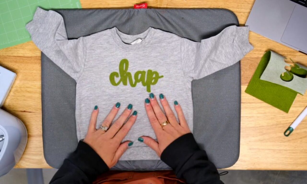 A pair of hands with teal nail polish and a ring align a gray, long-sleeved baby shirt on a pressing mat. The shirt features the word “chap” in green, with crafting supplies (including felt and a cutting mat) and a laptop visible in the background.