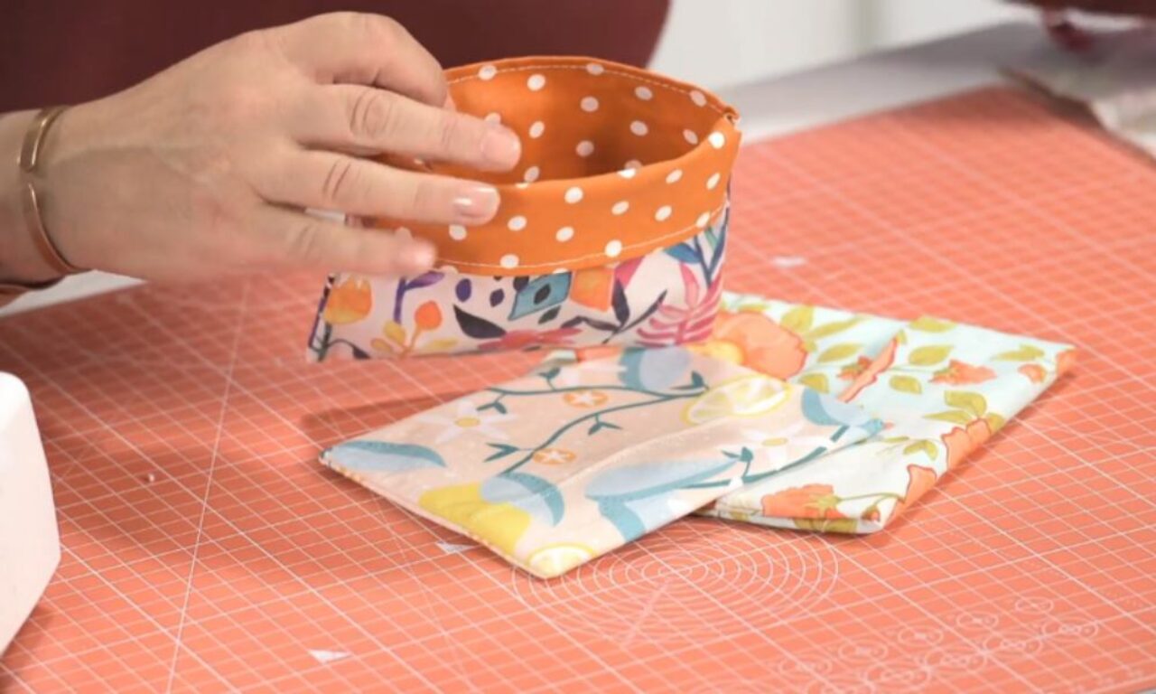 A hand holds an orange fabric pouch with white polka dots, while two colorful fabric pieces with floral patterns lie on a coral-colored cutting mat.