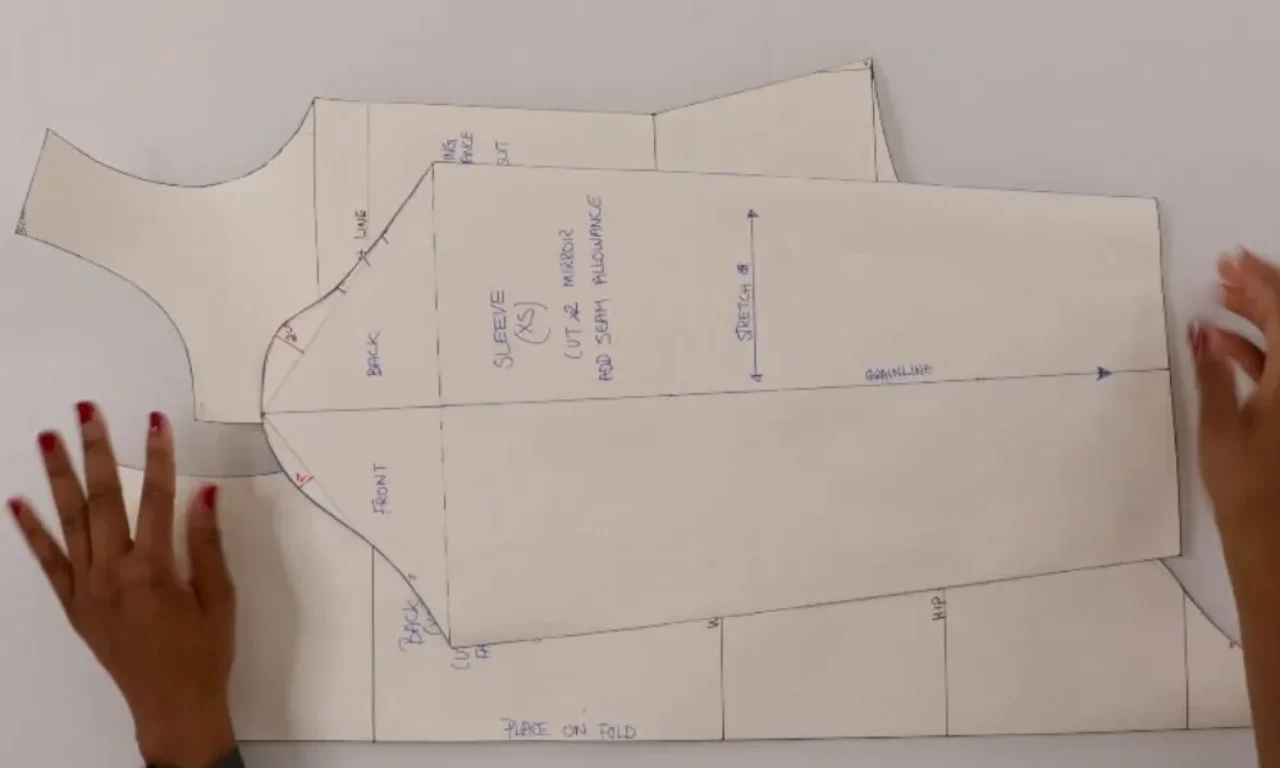 A pair of hands is arranging paper sewing patterns for a garment on a flat surface. The patterns are labeled with parts such as "Sleeve," "Front," and "Back," with instructions like "Cut 2 mirror" and "Add seam allowance." The patterns are neatly cut and include markings for grainline and stretch direction. The person has red nail polish on their fingernails.   
