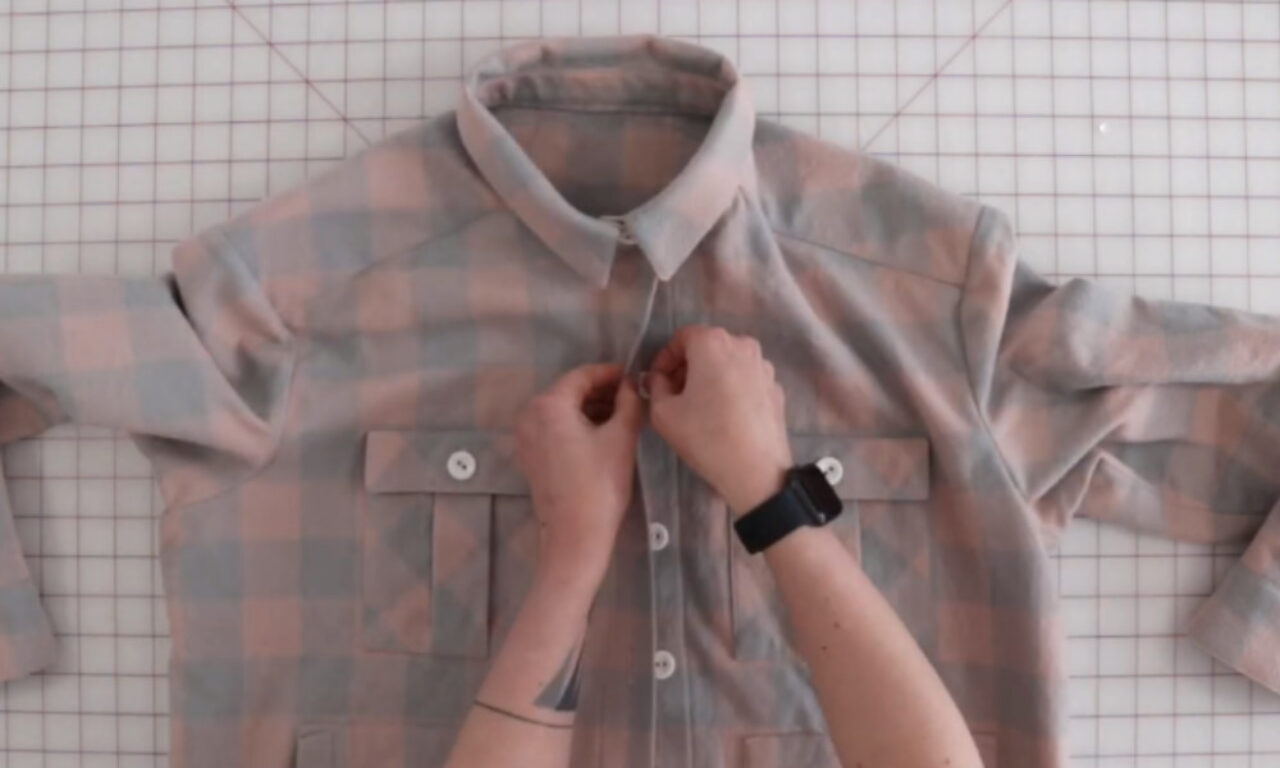 A person is buttoning up a pastel plaid shirt laid flat on a grid-patterned surface. The person's hands are visible, and they are wearing a black smartwatch on their left wrist.
