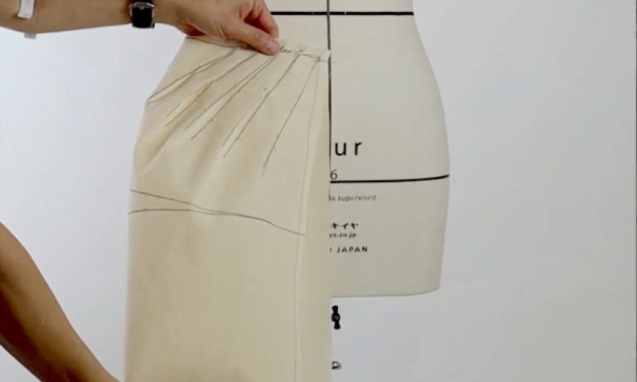 A close-up of a dressmaker's mannequin with fabric pinned and draped around the waist, showing pleats being created and arranged by a person's hand.