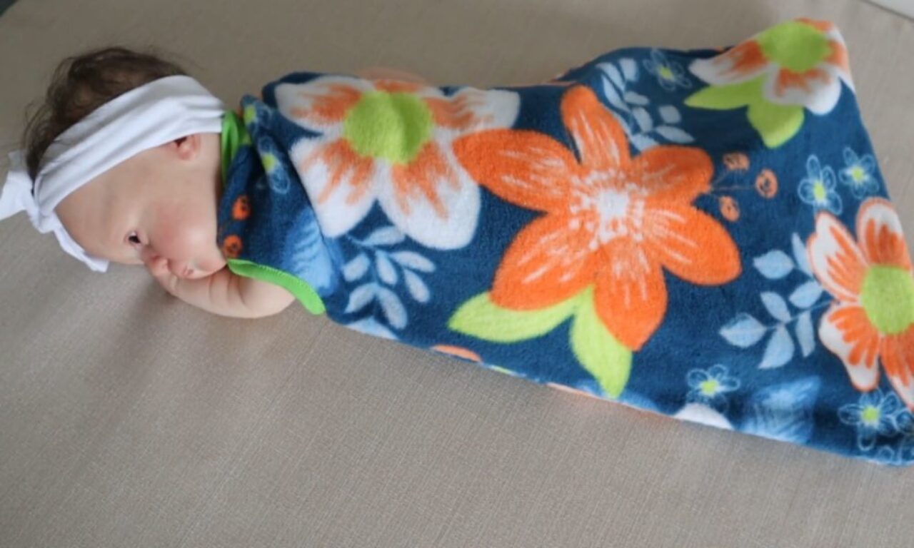 A baby lies on their side on a light-colored surface, swaddled in a soft blue fleece blanket decorated with large orange, white, and green flowers. The baby rests one hand near their face and wears a white headband tied in a bow.