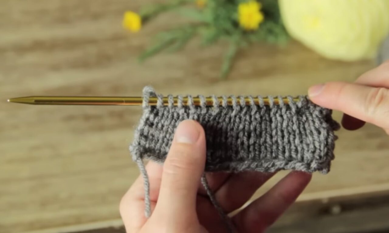 A close-up of a person's hands holding a small piece of gray knitted fabric on a gold knitting needle, with a ball of yarn and yellow flowers in the blurred background.