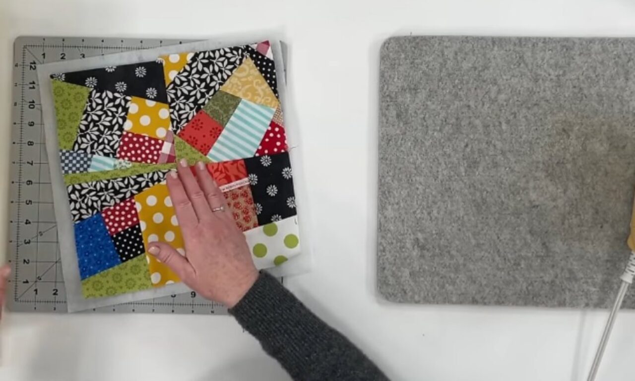 Top-down view of two hands straightening a colorful, scrappy quilt block on a gridded cutting mat; a gray wool pressing pad and the tip of a small iron sit to the right on the white workspace.