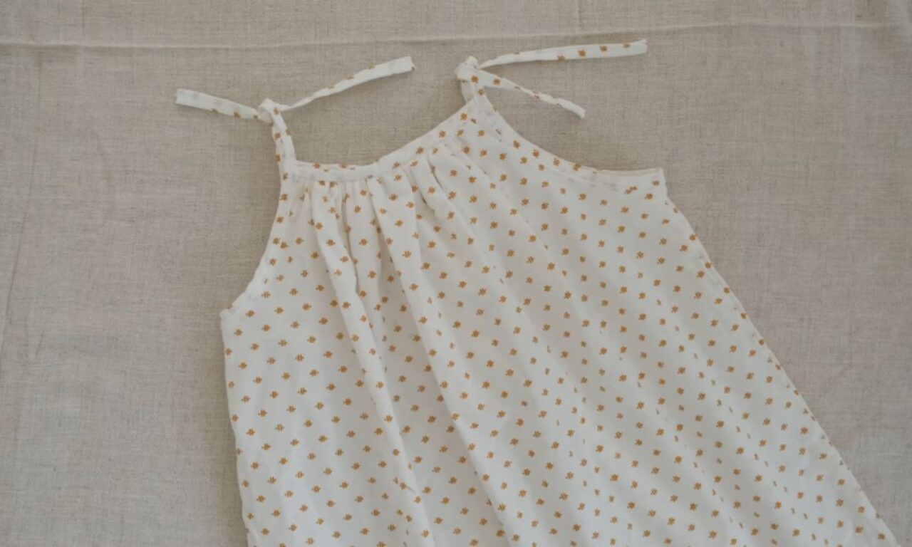 A sleeveless white cotton dress with small golden flower-shaped dots and shoulder ties, laid flat on a neutral linen background.