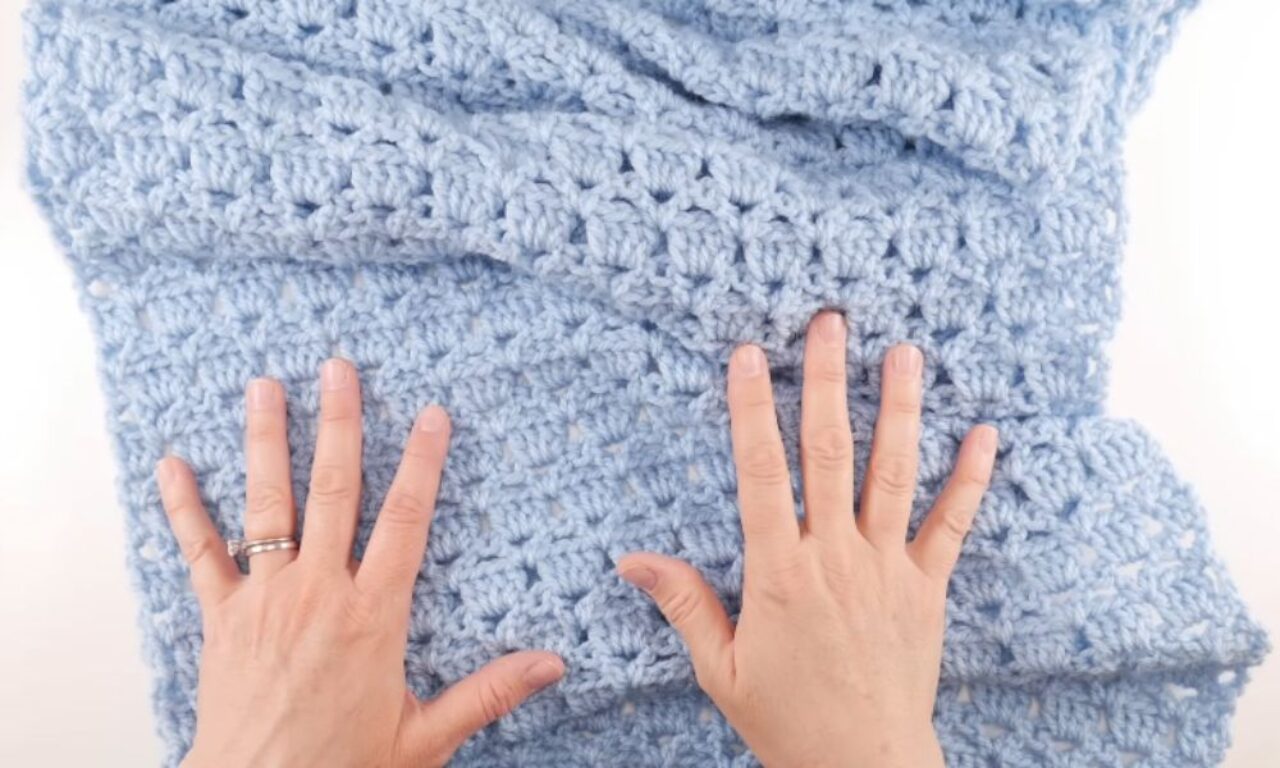 Hands gently press down on a light blue crochet blanket with a textured stitch pattern. One hand wears two rings.