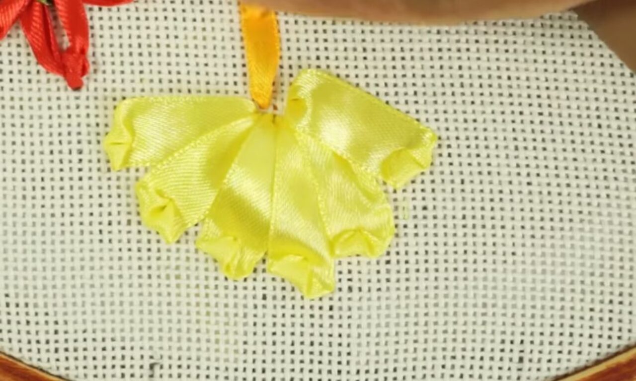 A close-up of a yellow satin ribbon folded and sewn onto a white woven fabric, forming a decorative petal-like pattern.