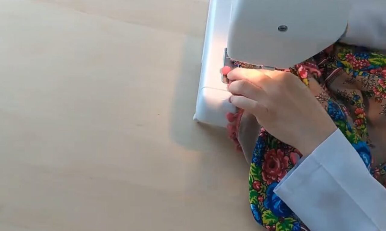 A person using a sewing machine to sew colorful fabric. The person's hand is guiding the fabric under the needle as the machine sews a straight line. The fabric has a vibrant floral pattern with red, blue, green, and yellow flowers. The sewing machine is white and sits on a light-colored wooden table. The person wears a white long-sleeved shirt.