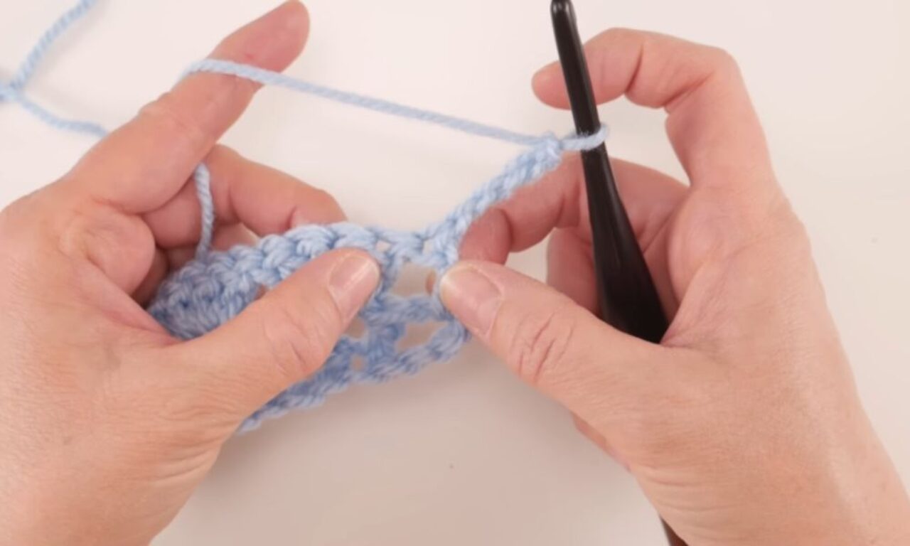 Close-up of two hands crocheting with light blue yarn and a black crochet hook. The left hand is holding the crocheted fabric, while the right hand holds the hook with yarn wrapped around it, in the process of making a stitch. The background is a plain white surface.