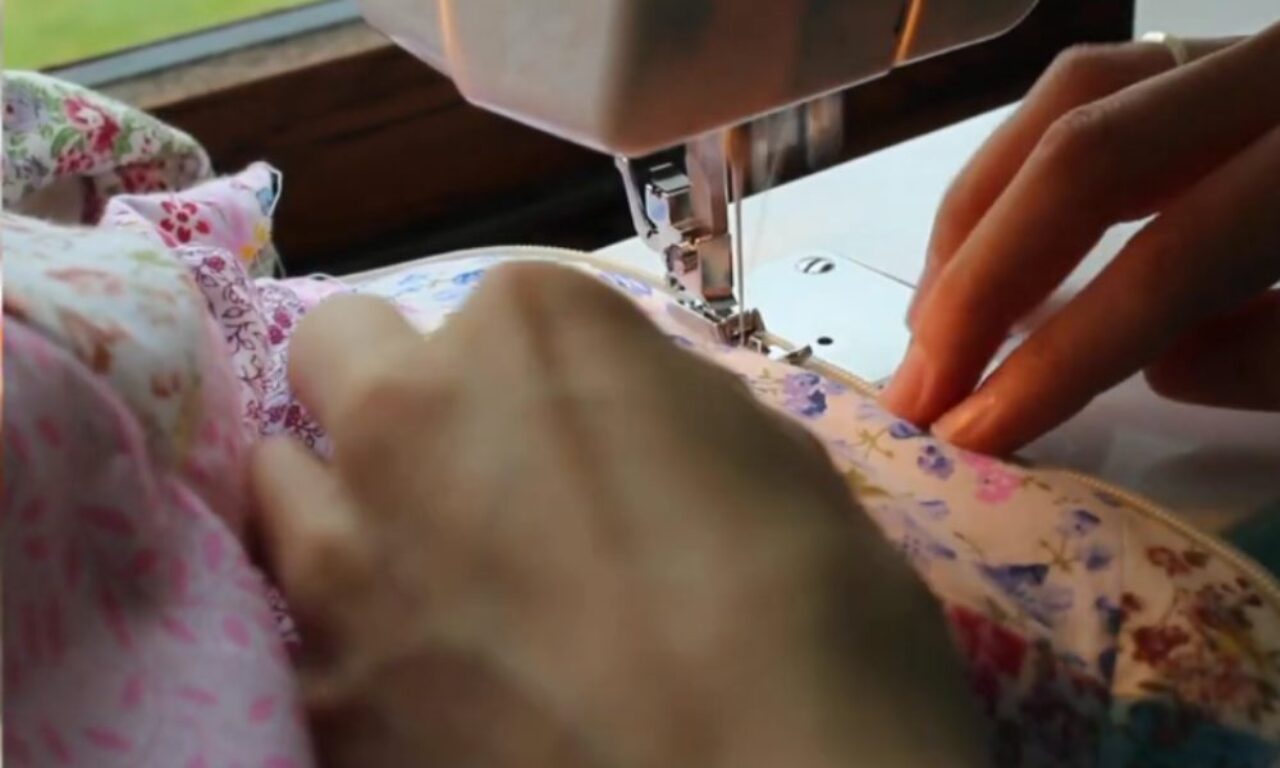 hands sewing a colorful floral fabric on a white sewing machine, with a window revealing a green outdoor scene in the background.