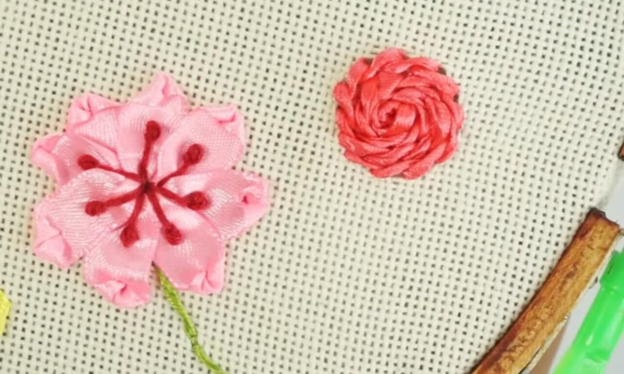 Two handcrafted fabric flowers on a textured background. The larger flower on the left is pink with a star-like center and ruffled petals. The smaller flower on the right is also pink and has a tightly wound, spiral shape. A pair of scissors with green and brown handles is partially visible in the bottom right corner of the image.