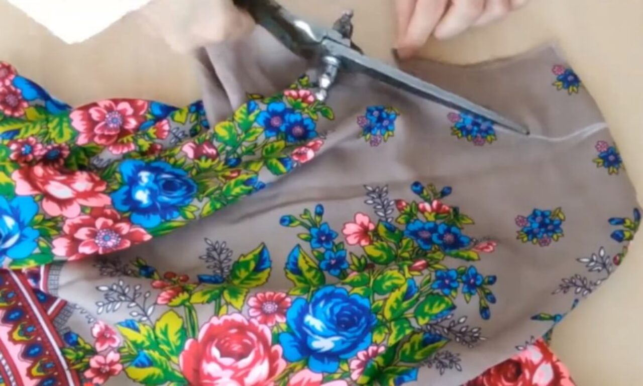 A person's hands holding a pair of scissors and cutting a piece of fabric with a colorful floral pattern. The fabric is primarily gray with blue, pink, and red flowers and green leaves.