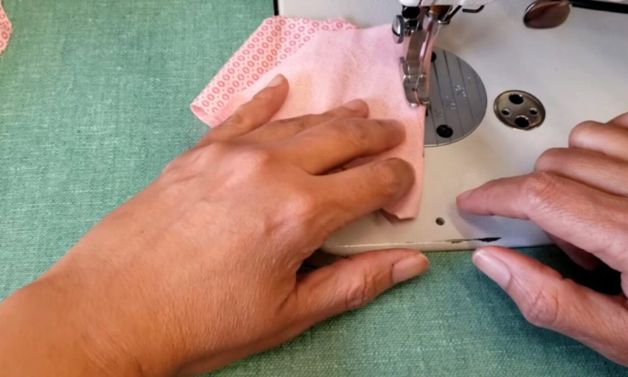 A person is sewing a piece of pink fabric on a white sewing machine. Their hands are guiding the fabric under the needle, with one hand holding the fabric in place and the other adjusting the machine. The background is a green surface, likely a table or work mat.