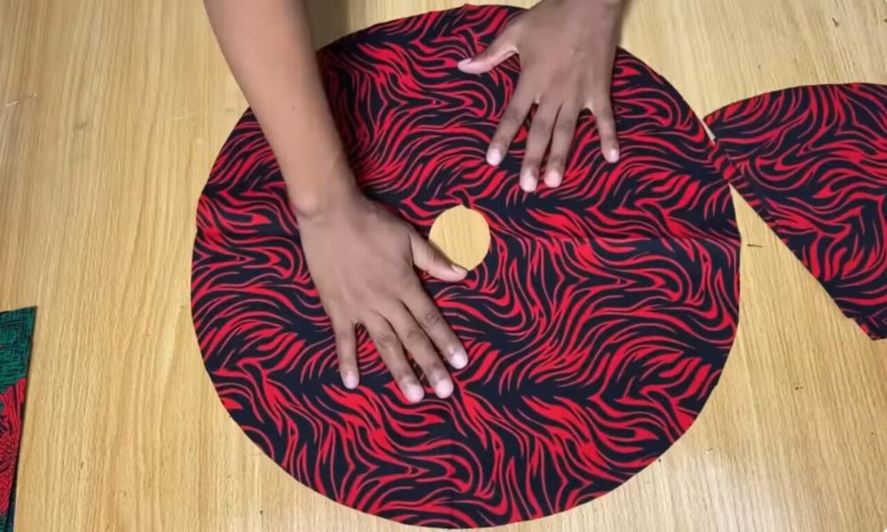A person's hands are shown working on a circular piece of red and black patterned fabric on a wooden table. The fabric is cut into a spiral shape and a round wooden object is visible in the center of the spiral.