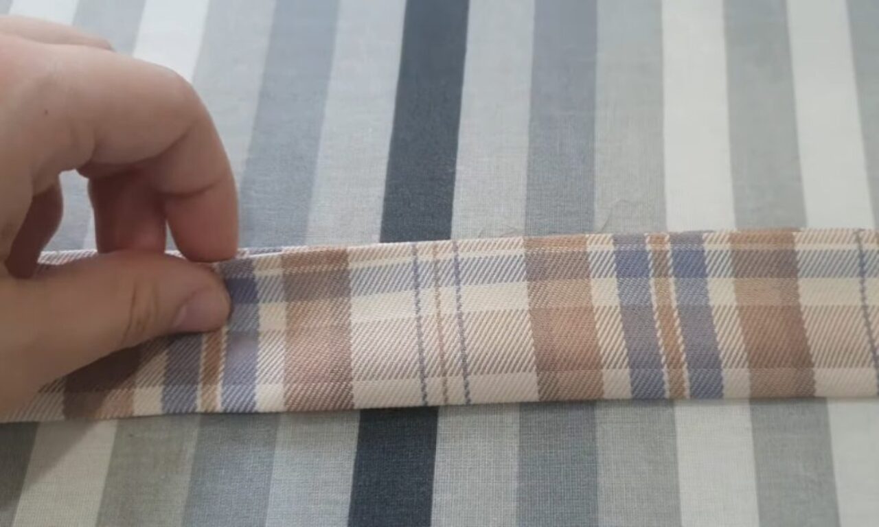 A close-up of a hand rolling up a piece of fabric with a blue, brown, and white plaid pattern, on top of a larger piece of fabric with blue, gray, and white stripes.