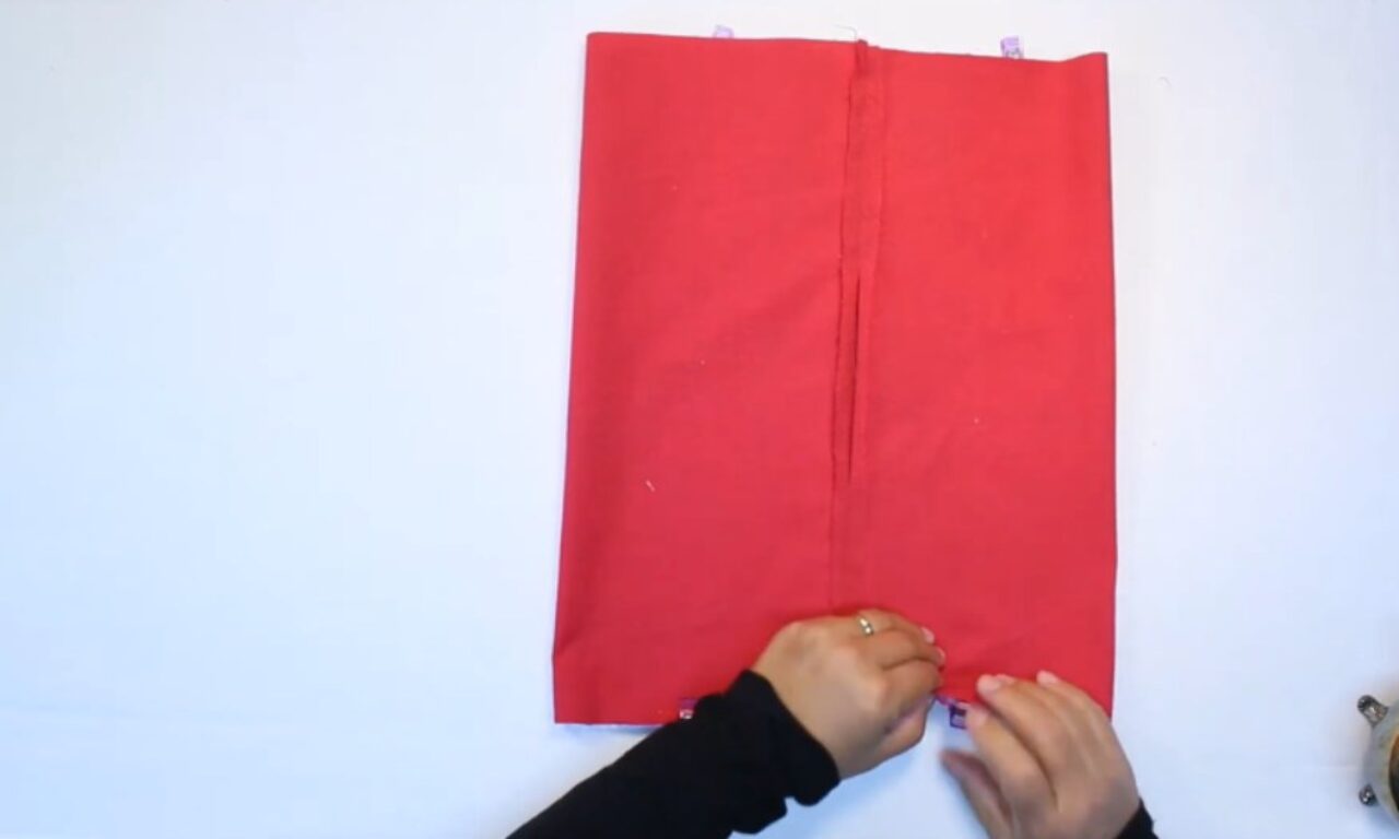 An overhead view of hands on a white surface, carefully securing the bottom edge of a large red fabric rectangle with small purple clips. The fabric has a prominent vertical seam running down its center. Additional clips are visible along the top edge.