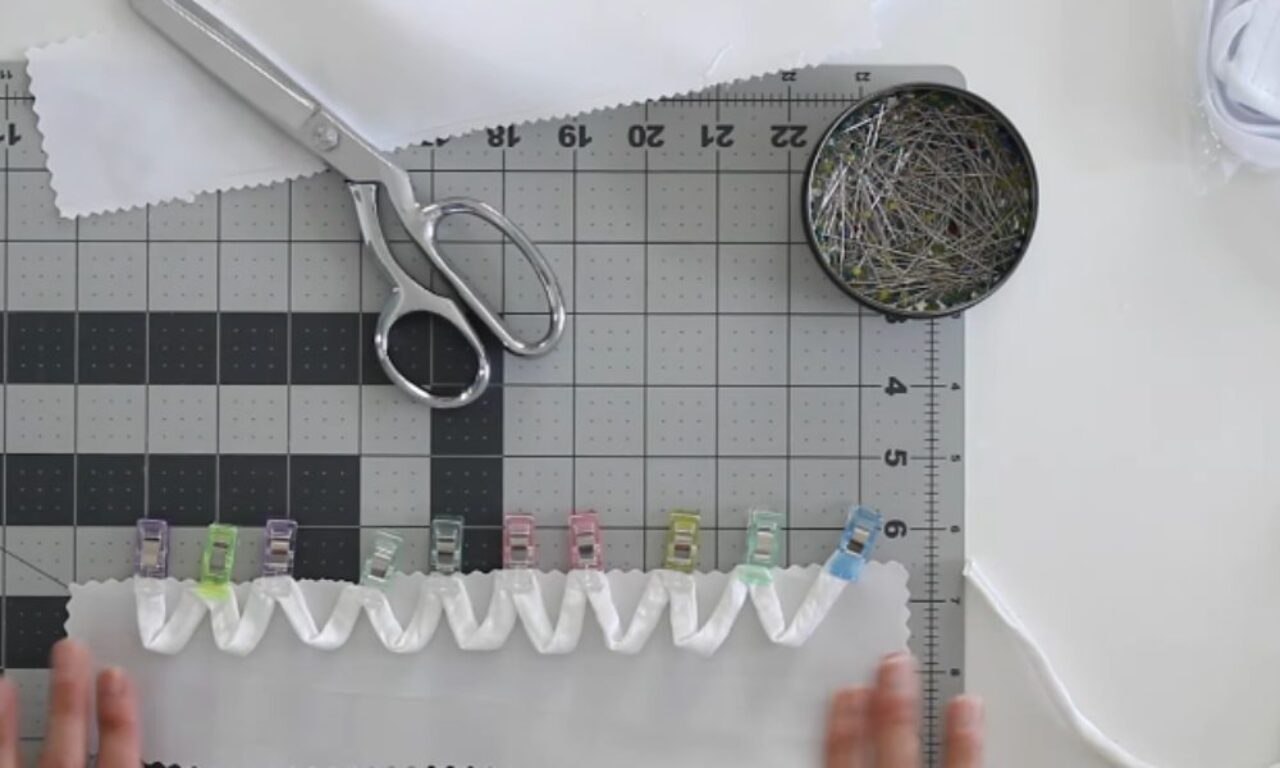 An overhead view of a sewing project on a grey cutting mat. A strip of white elastic is laid in a wavy pattern across a piece of white fabric and secured with colorful sewing clips. Silver pinking shears and a tin of sewing pins are at the top, and a person's hands are visible holding the fabric at the bottom.