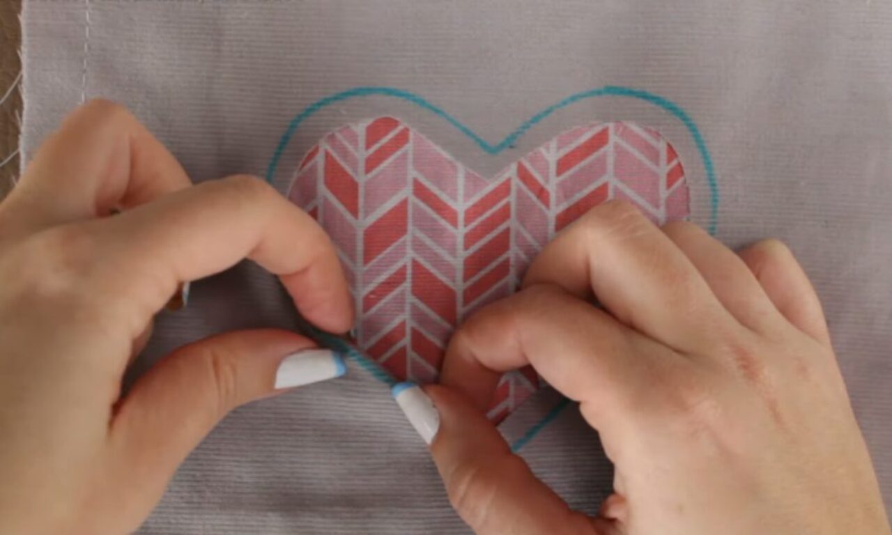 Hands holding a heart-shaped fabric piece with a red and white chevron pattern, positioned on a light gray fabric background. The heart is outlined with a blue marker, suggesting a sewing or crafting project in progress.