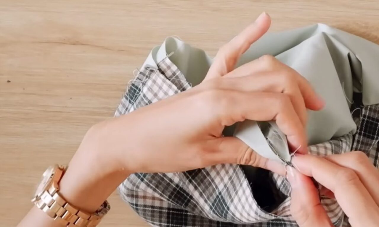 Hands stitching fabric with a needle and thread on a wooden surface. One hand is holding the fabric while the other is guiding the needle through the material. The person is wearing a gold watch on their wrist. The fabric includes a checkered pattern and a solid light green section.