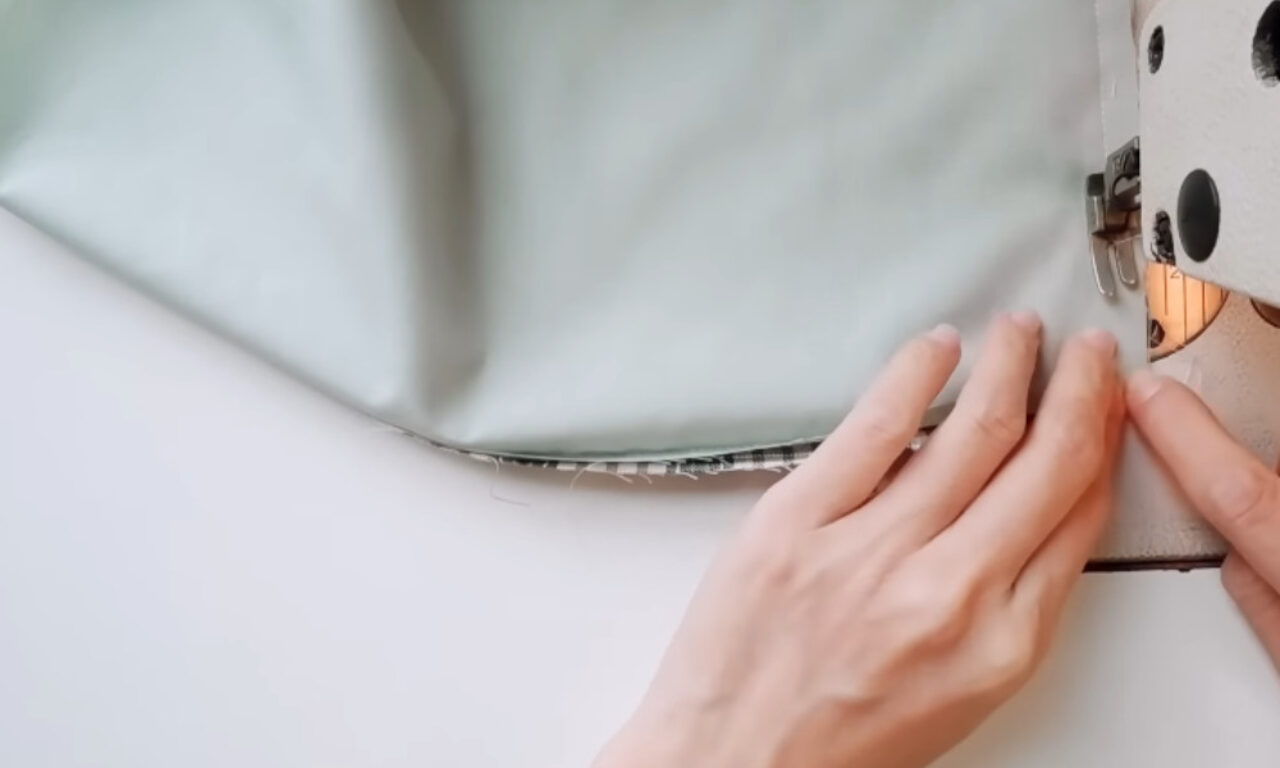 Hands guide light green fabric through a sewing machine.