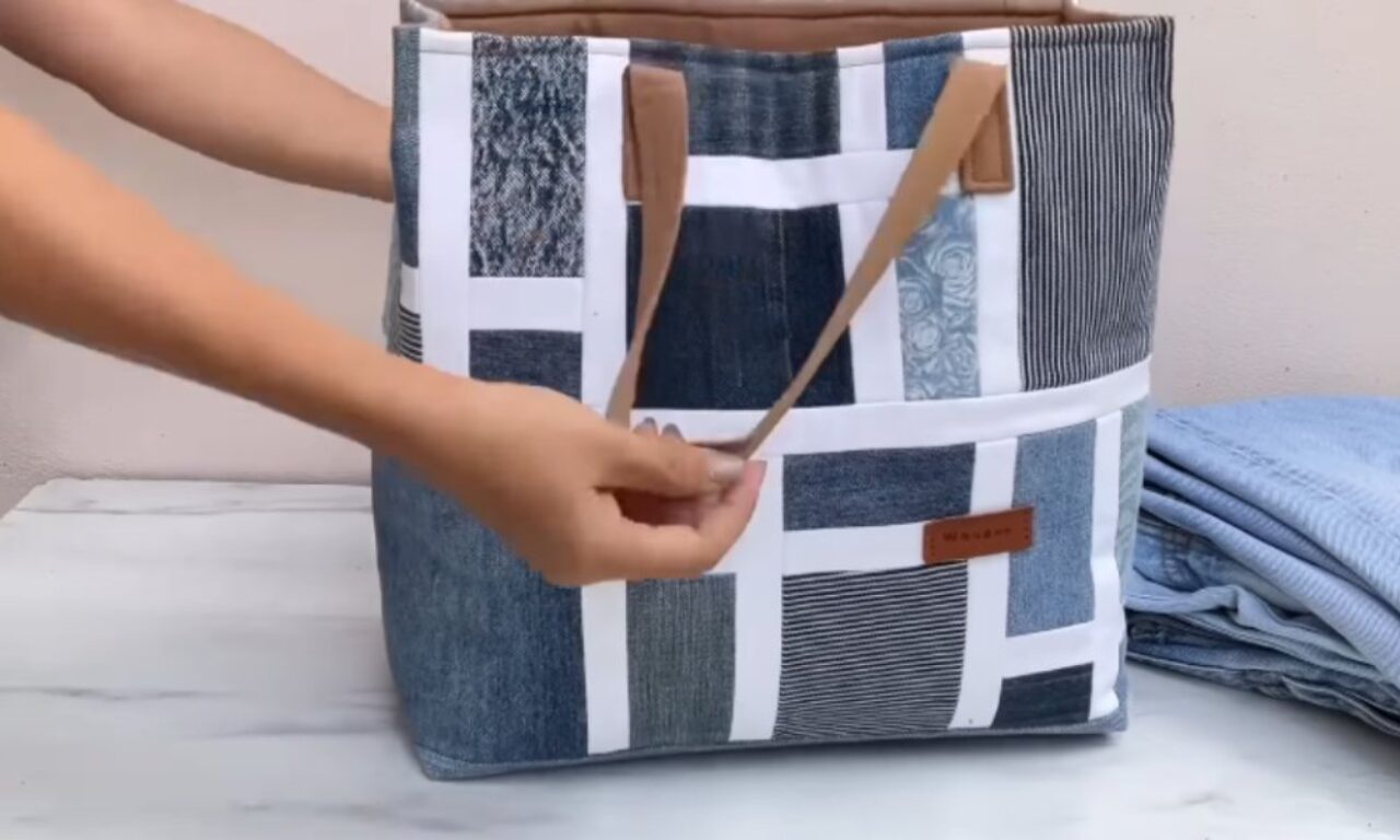 a handcrafted tote bag, featuring a patchwork design of various denim washes and textures sewn together with white seams. Brown handles are held by a hand. The bag is on a marble surface with folded denim pants visible in the background.