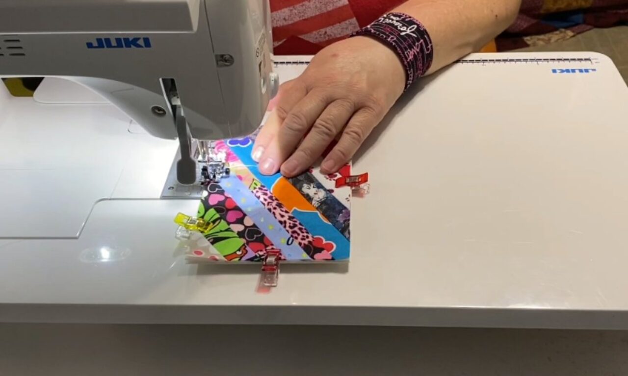 A person's right hand guides a vibrant patchwork fabric under the needle of a white JUKI sewing machine. The fabric is a patchwork of diagonal strips in various colorful patterns, held together by several colorful sewing clips. The person wears a dark wristband with pink text on their right wrist.