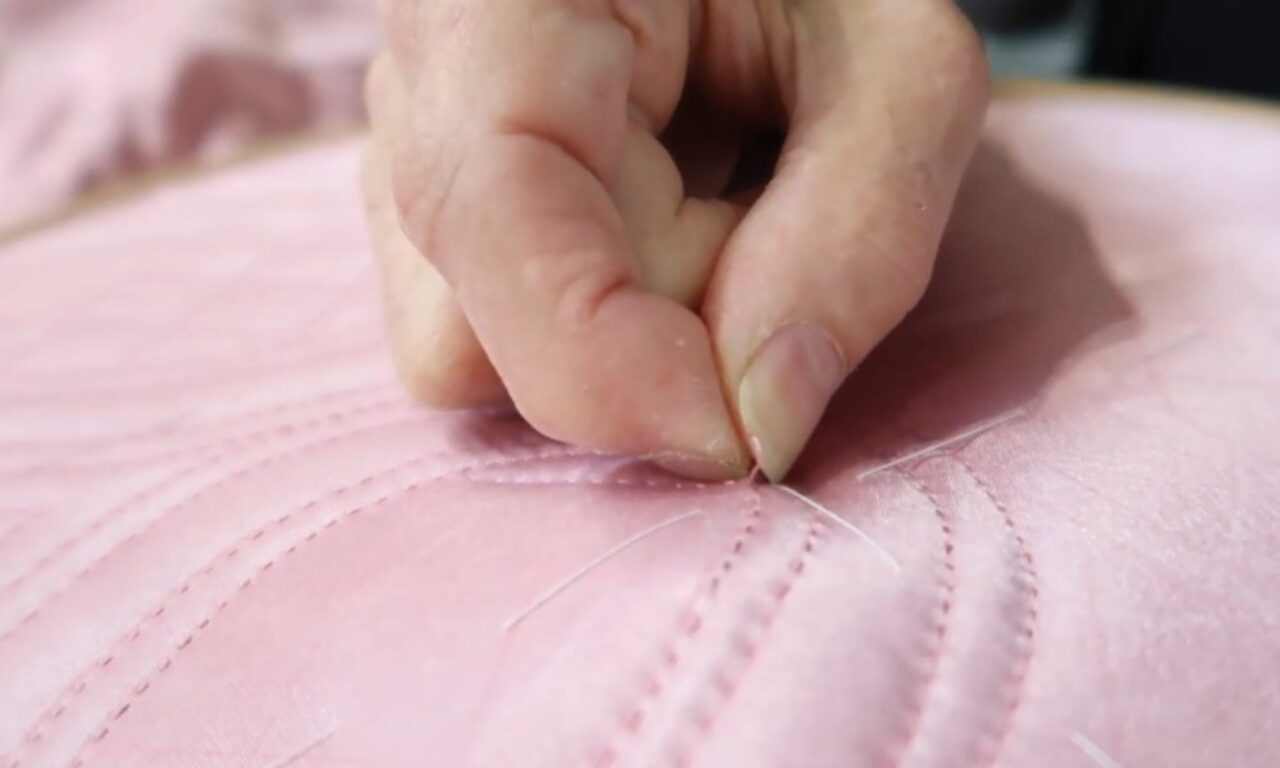 A close-up of a hand stitching a pink fabric, the needle and thread creating a delicate pattern. The hand moves in and out of frame as it sews, the fabric smooth and slightly shiny.