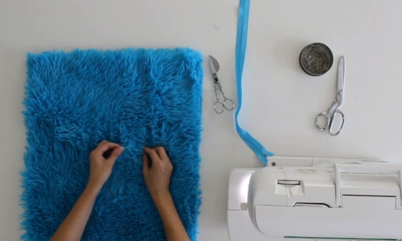 A pair of hands working with a piece of bright blue faux fur fabric on a white table. To the right, there are sewing tools including a pair of scissors, a small bowl of pins, a seam ripper, a strip of blue fabric or ribbon, and a sewing machine.