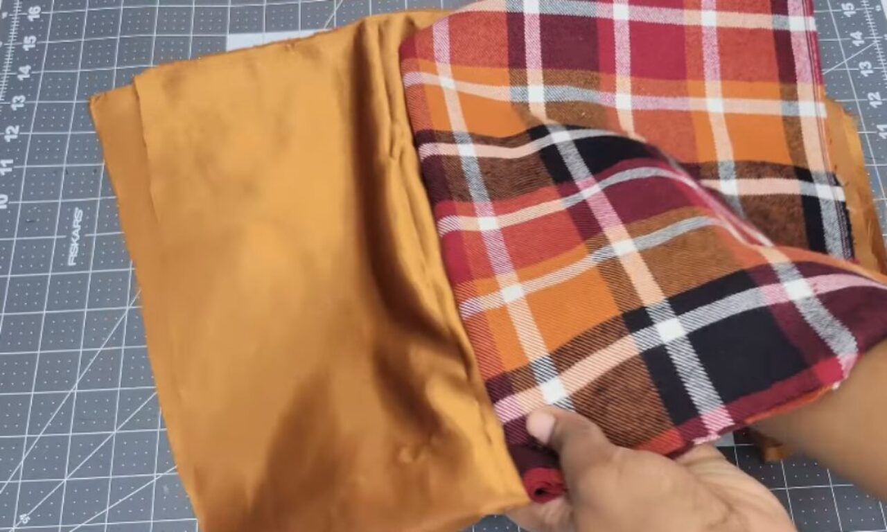 A person's hands are holding two pieces of fabric on a cutting mat. The fabric on the left is a solid mustard yellow, while the fabric on the right is a plaid pattern with red, orange, brown, and white colors. The cutting mat has a grid pattern for precise measurements.