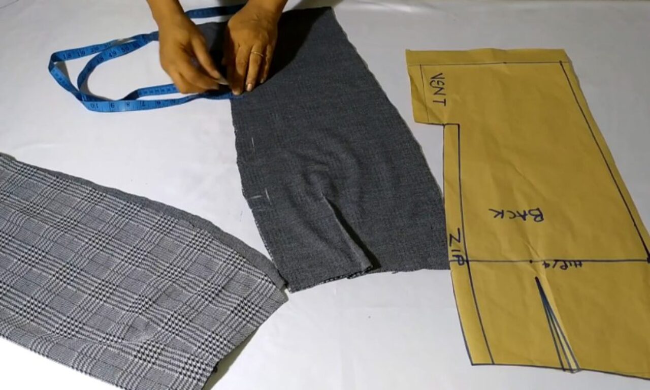 A person is working on a sewing project. They are using a measuring tape to adjust a piece of dark gray fabric, which appears to be part of a garment. Next to the gray fabric, there is a piece of checkered fabric and a yellow paper pattern labeled with sewing instructions and markings such as 'VENT' and 'BACK.' The scene suggests the process of tailoring or garment construction.