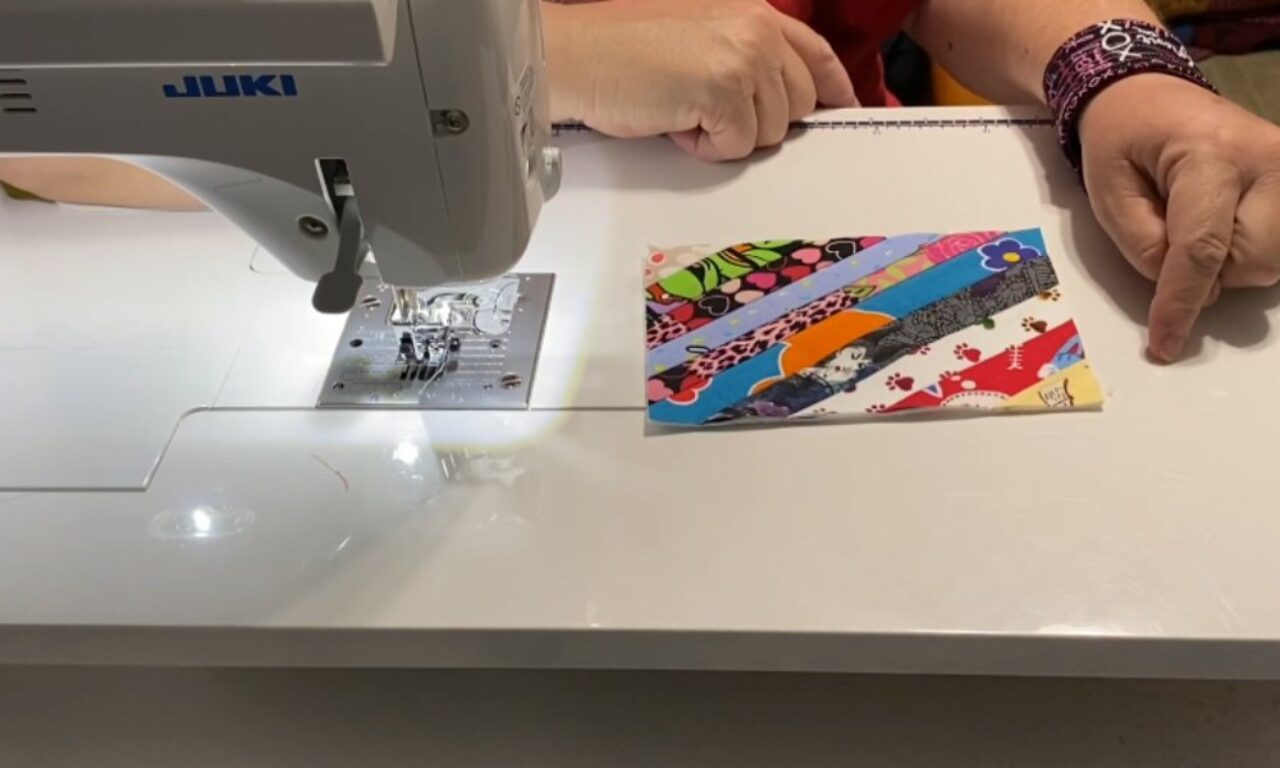 A person sits at a sewing machine, with their hands resting on the table. In front of them is a small rectangular piece of fabric made from colorful, patterned strips sewn together. The sewing machine is white and labeled “JUKI.” The person wears a purple patterned wristband on their right wrist.