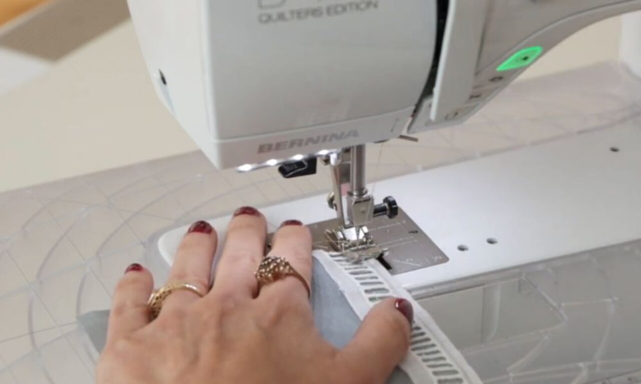 a person's hand guiding fabric on a white Bernina Quilter's Edition sewing machine. The hand, with red nail polish and rings, holds a piece of gray fabric as a white, ribbed trim is stitched to its edge under the needle. The machine's bright LED light illuminates the work area, which sits on a clear, gridded extension table.