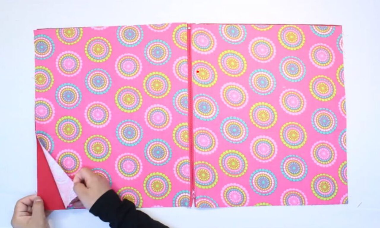 A person's hands are folding a piece of colorful wrapping paper with a pink background and circular patterns in various bright colors. The paper is being folded along a crease to create a neat edge.