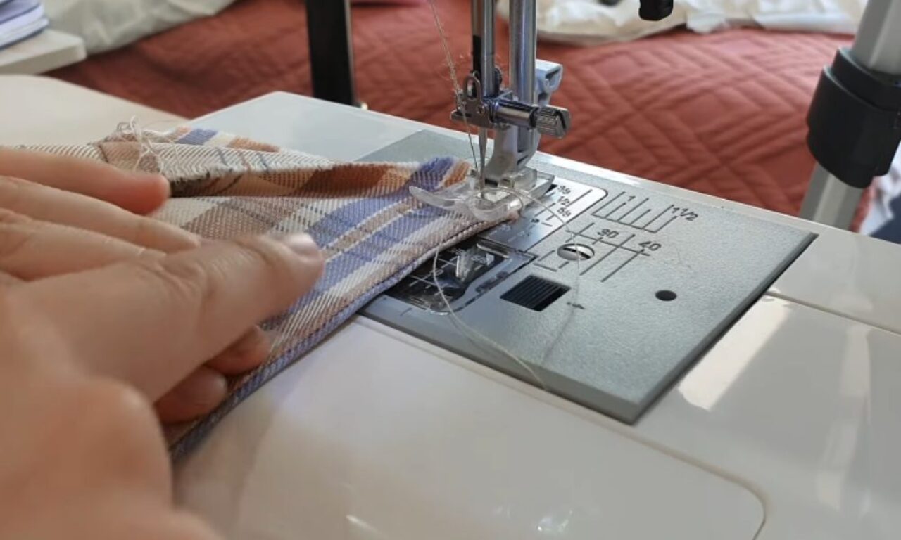 A person is sewing a striped fabric on a white sewing machine. The fabric is being guided under the needle by the person's hands, and the machine is positioned on a table or surface in front of a bed with a red quilted comforter.