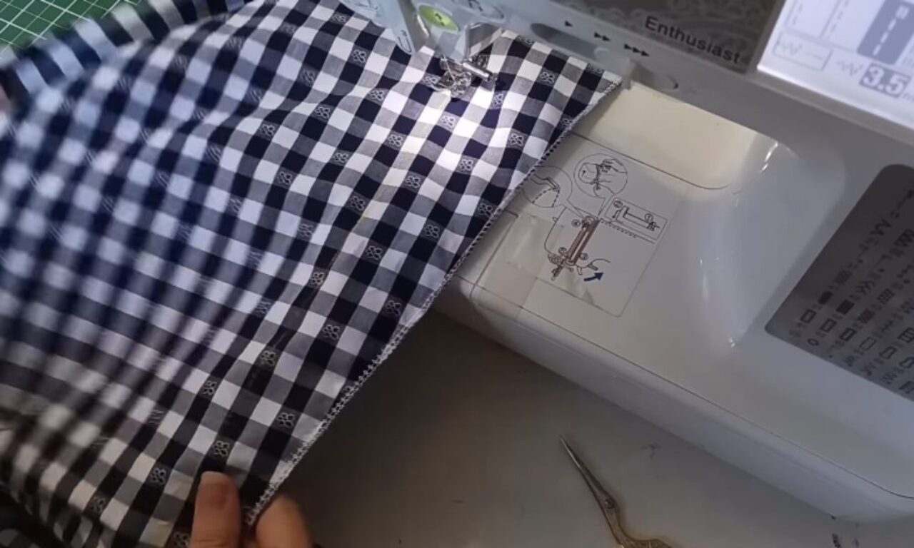 A person is using a sewing machine to stitch the edge of a piece of black and white checkered fabric. The fabric is being guided under the presser foot, and a pair of small embroidery scissors lies nearby on the table.