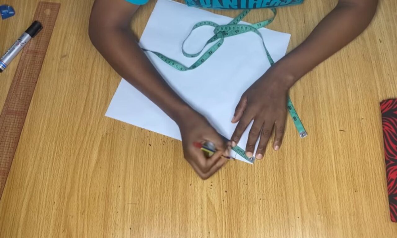Overhead view of a person marking measurements on a white sheet of paper with a pencil, using a green sewing tape measure on a wooden worktable; a clear ruler and marker rest nearby.