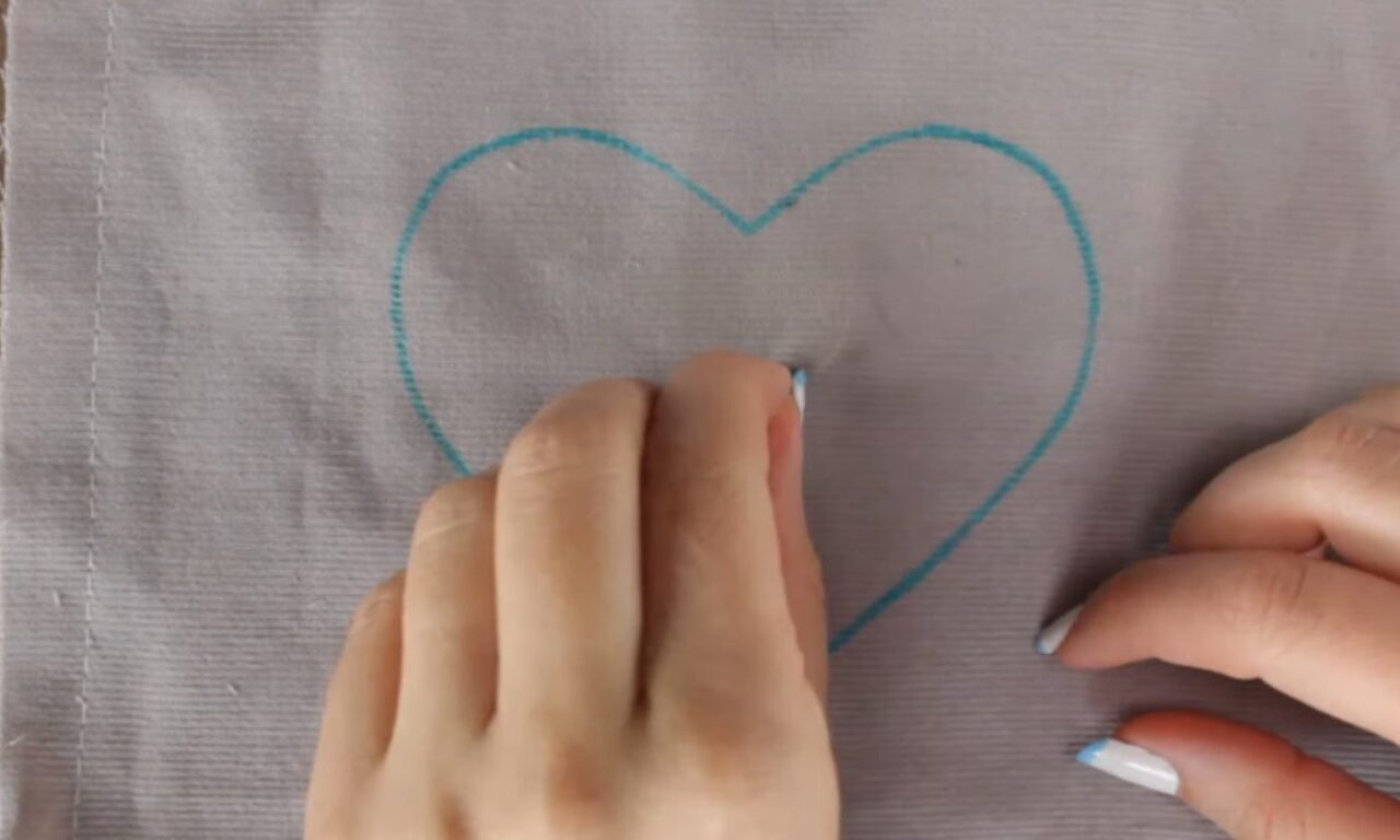 Two hands drawing a heart shape with a blue marker on a piece of light-colored fabric.
