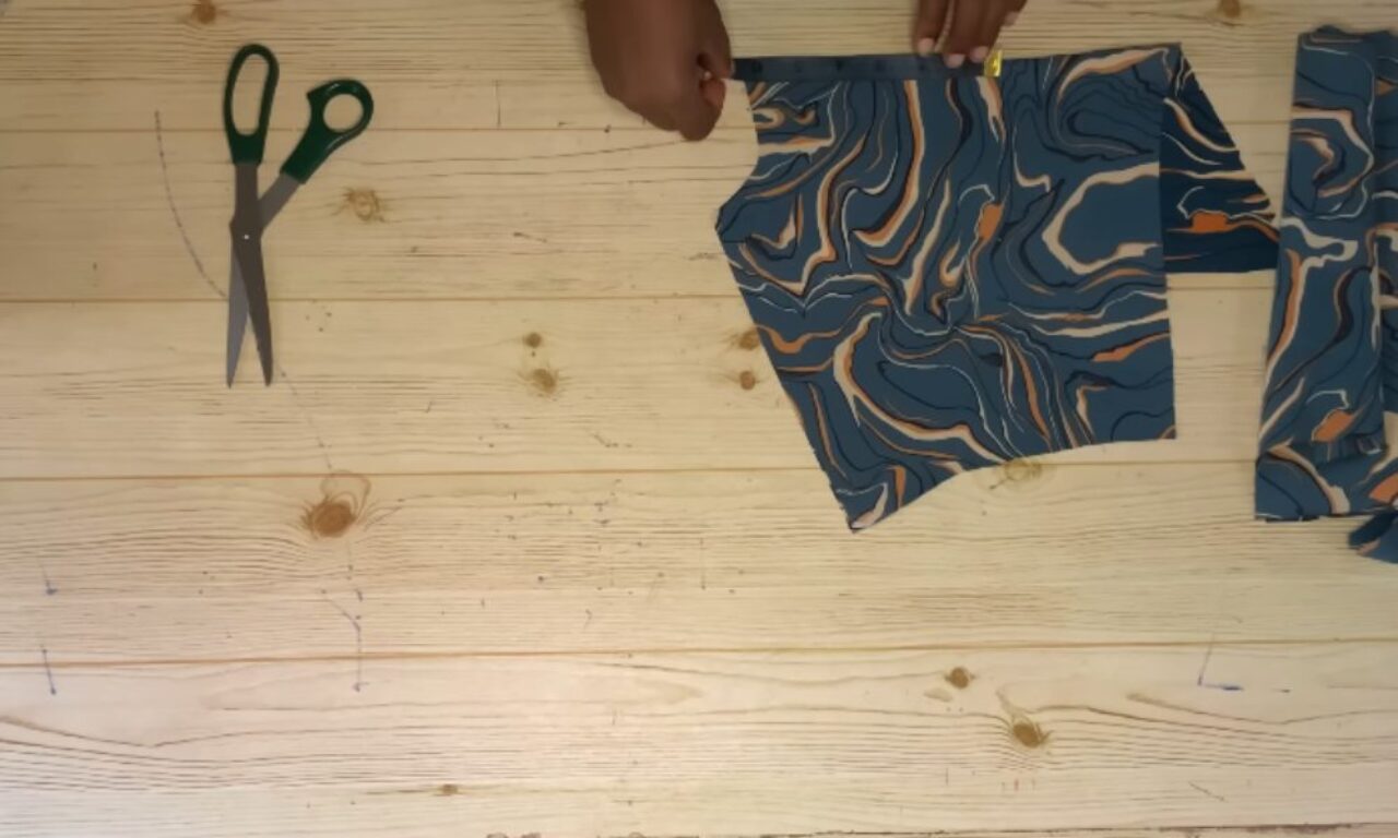 The image shows a pair of green-handled scissors placed on a light wooden surface. Next to the scissors, there are two pieces of fabric with a blue and orange abstract pattern. A person's hand is visible, holding one of the fabric pieces, suggesting they are in the process of cutting or arranging the fabric. The scene appears to be part of a sewing or crafting project.