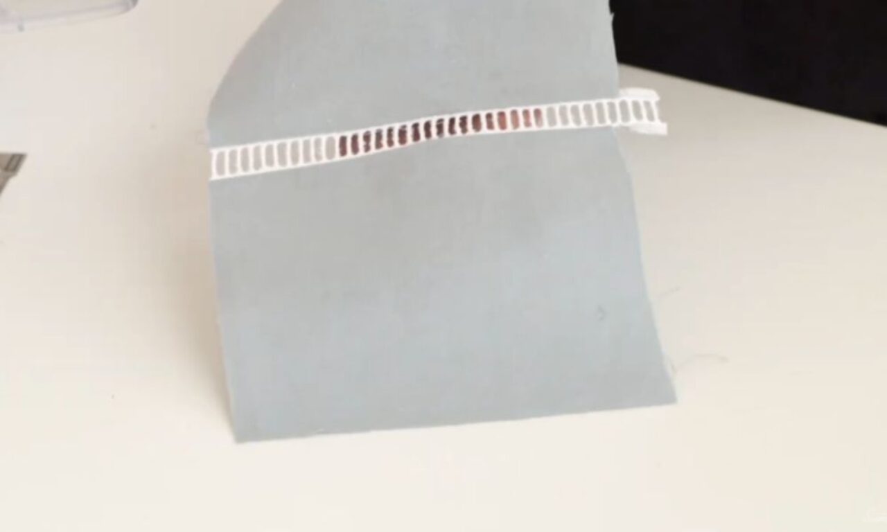 A small strip of white plastic ladder-like material or tape with reddish-brown markings is placed across a folded gray paper or card. The items are sitting on a white surface, with a dark background visible at the top right of the image. This appears to be some kind of craft, laboratory sample, or testing material setup.