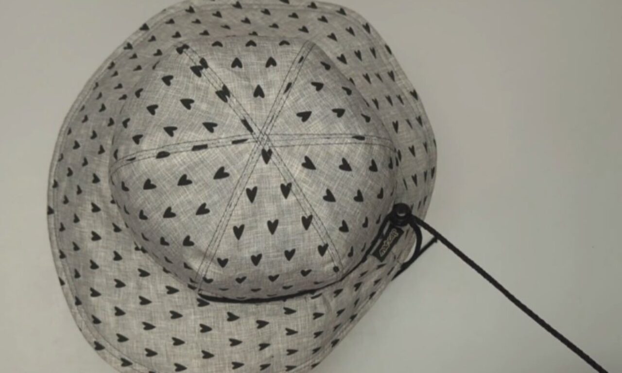 A light grey bucket hat patterned with numerous small black hearts. The hat features a segmented crown with visible stitching and a wide brim. A black chin strap with a toggle and a black brand tag, possibly reading "ROKKA," is attached to one side of the brim. The hat is laid flat against a plain, light background.